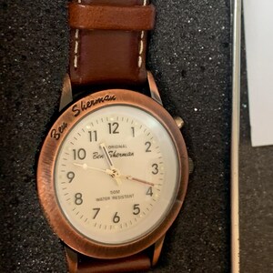 K&ouml;nnte beinhalten: Eine Ben Sherman Uhr mit einem braunen Lederarmband und einem kupferfarbenen Geh&auml;use. Das wei&szlig;e Zifferblatt zeigt schwarze Ziffern und Zeiger. Der Text "Ben Sherman" und "50M Water Resistant" sind sichtbar. Die Uhr ist wasserdicht.