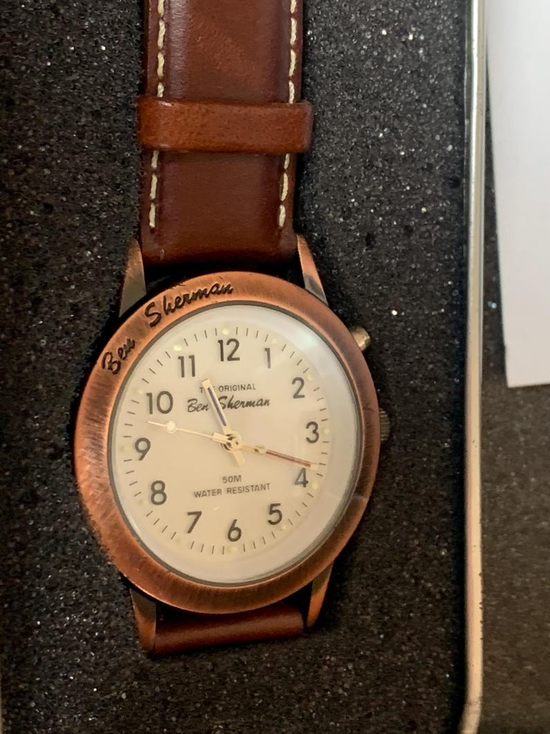 K&ouml;nnte beinhalten: Eine Ben Sherman Uhr mit einem braunen Lederarmband und einem kupferfarbenen Geh&auml;use. Das wei&szlig;e Zifferblatt zeigt schwarze Ziffern und Zeiger. Der Text "Ben Sherman" und "50M Water Resistant" sind sichtbar. Die Uhr ist wasserdicht.