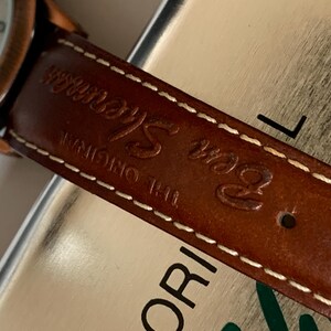 K&ouml;nnte beinhalten: Nahaufnahme eines braunen Leder-Uhrenarmbands mit wei&szlig;en N&auml;hten und dem gepr&auml;gten Text "Ben Sherman THE ORIGINAL". Das Zifferblatt der Uhr ist sichtbar, mit einer kupferfarbenen L&uuml;nette und einem wei&szlig;en Zifferblatt.