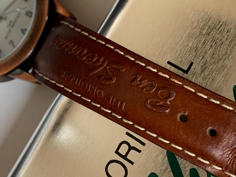 K&ouml;nnte beinhalten: Nahaufnahme eines braunen Leder-Uhrenarmbands mit wei&szlig;en N&auml;hten und dem gepr&auml;gten Text "Ben Sherman THE ORIGINAL". Das Zifferblatt der Uhr ist sichtbar, mit einer kupferfarbenen L&uuml;nette und einem wei&szlig;en Zifferblatt.