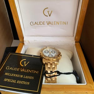 Könnte beinhalten: Eine goldfarbene Claude Valentini Millennium Ladies Special Edition Uhr mit weißem Zifferblatt und drei Hilfszifferblättern, präsentiert in einer Holzschatulle mit einem schwarzen und goldenen Etikett mit Markenlogo und Modellname.