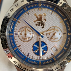 Puede incluir: Reloj de pulsera de acero inoxidable con esfera blanca, detalles azules y dorados, y un patr&oacute;n texturizado. El reloj presenta un emblema de le&oacute;n escoc&eacute;s, subesferas y el texto "Scotland Forever".