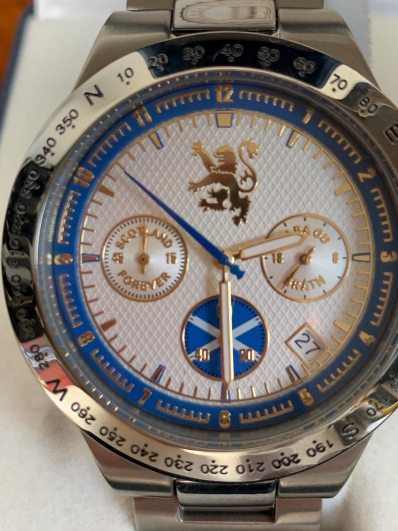 Puede incluir: Reloj de pulsera de acero inoxidable con esfera blanca, detalles azules y dorados, y un patr&oacute;n texturizado. El reloj presenta un emblema de le&oacute;n escoc&eacute;s, subesferas y el texto "Scotland Forever".
