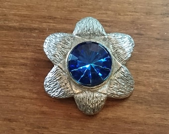 May include: A silver flower-shaped brooch with a large blue gemstone in the center.