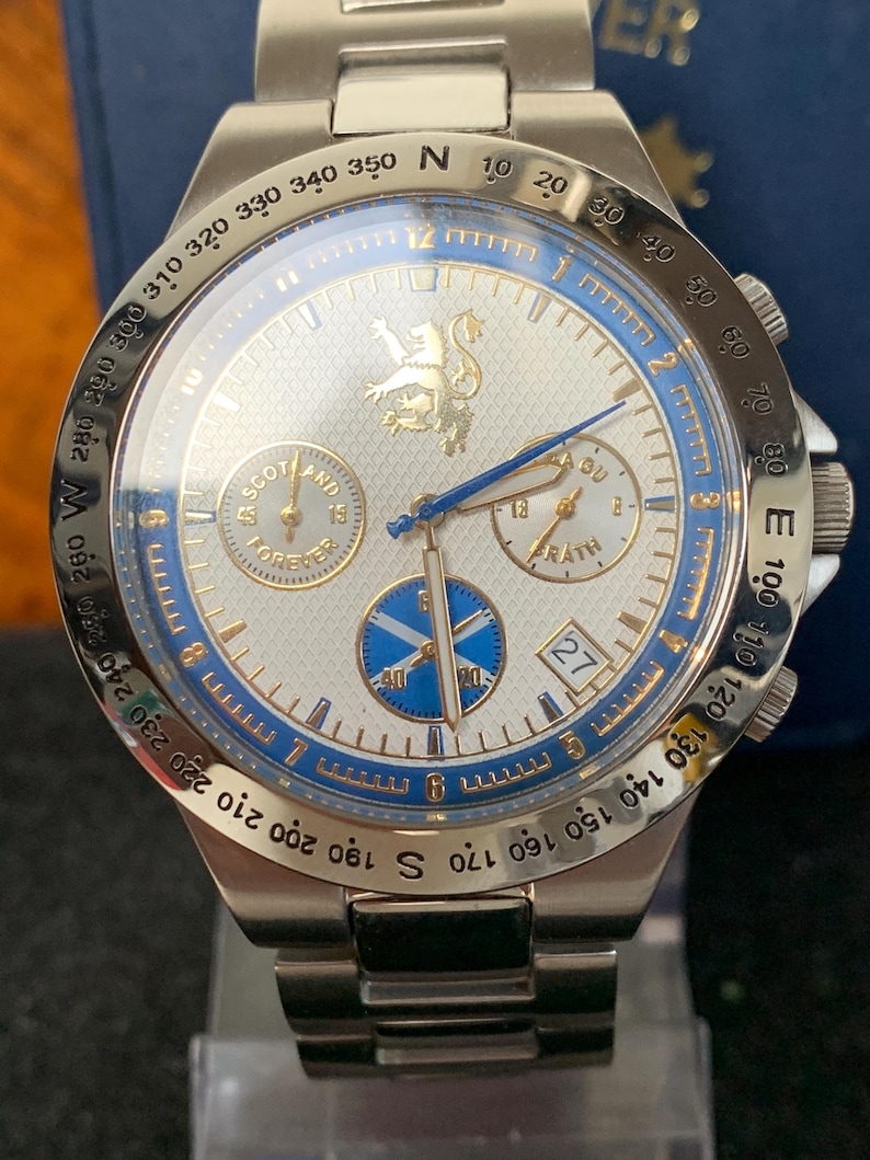 Puede incluir: Un reloj de pulsera plateado con esfera blanca, detalles azules y correa de acero inoxidable. El reloj presenta un emblema dorado y el texto "SCOTLAND FOREVER". Incluye una pantalla de fecha y varias subesferas.