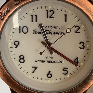 K&ouml;nnte beinhalten: Nahaufnahme einer Ben Sherman Uhr mit cremefarbenem Zifferblatt und kupferfarbener L&uuml;nette. Das Zifferblatt zeigt schwarze Ziffern, den Text "THE ORIGINAL Ben Sherman" und "50M WATER RESISTANT". Das Armband ist aus braunem Leder.