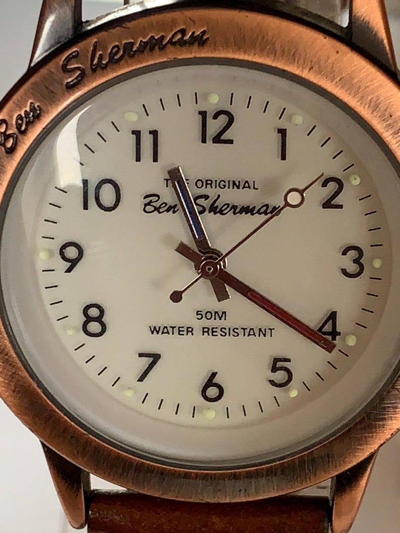 K&ouml;nnte beinhalten: Nahaufnahme einer Ben Sherman Uhr mit cremefarbenem Zifferblatt und kupferfarbener L&uuml;nette. Das Zifferblatt zeigt schwarze Ziffern, den Text "THE ORIGINAL Ben Sherman" und "50M WATER RESISTANT". Das Armband ist aus braunem Leder.