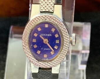 May include: A vintage Sekonda wristwatch with a silver-tone metal band and case. The watch face is a deep blue with gold-tone hands and markers. The watch reads "SEKONDA" and "17 jewels". The watch is displayed on a clear stand.