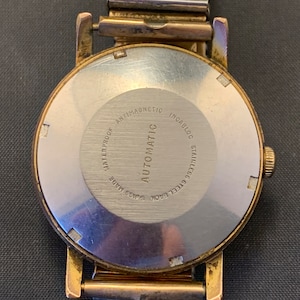May include: Close-up of a vintage gold-toned wristwatch with a silver-colored back. The watch back is engraved with text including "WATERPROOF," "ANTIMAGNETIC," and "STAINLESS STEEL BACK." The watch band is a silver-colored Fixo-Flex band.