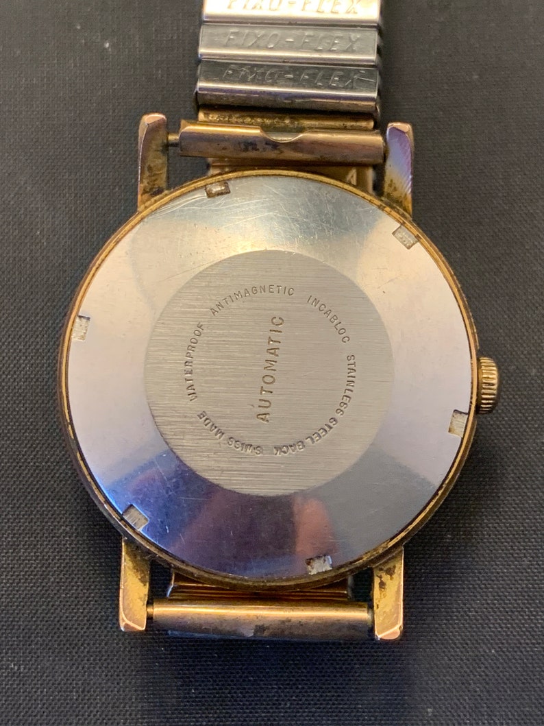 May include: Close-up of a vintage gold-toned wristwatch with a silver-colored back. The watch back is engraved with text including "WATERPROOF," "ANTIMAGNETIC," and "STAINLESS STEEL BACK." The watch band is a silver-colored Fixo-Flex band.