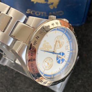 Puede incluir: Un reloj de pulsera plateado con esfera blanca y detalles azules. El reloj presenta detalles dorados, incluido un logotipo y n&uacute;meros. La correa del reloj es de metal plateado. El reloj est&aacute; sobre un fondo azul con la palabra "SCOTLAND".