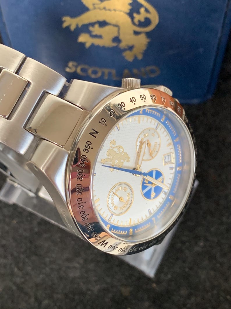 Puede incluir: Un reloj de pulsera plateado con esfera blanca y detalles azules. El reloj presenta detalles dorados, incluido un logotipo y n&uacute;meros. La correa del reloj es de metal plateado. El reloj est&aacute; sobre un fondo azul con la palabra "SCOTLAND".