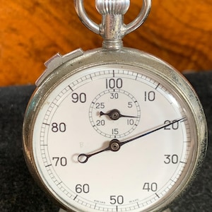 Op de afbeelding: Een zilveren zakhorloge met een witte wijzerplaat en zwarte wijzers. Het horloge heeft een ring aan de bovenkant en staat op een standaard. De wijzerplaat van het horloge heeft cijfers van 10 tot 100, en de woorden "Swiss Made" staan ​​onderaan gedrukt.