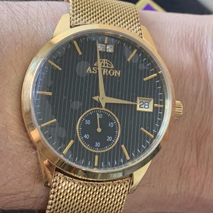 May include: A gold-tone wristwatch with a black face and a mesh band. The watch face has a small date window at the 3 o'clock position and the word "Astron" printed on it.