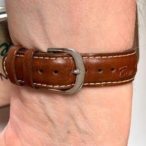 K&ouml;nnte beinhalten: Nahaufnahme eines braunen Leder-Uhrenarmbands mit wei&szlig;en N&auml;hten und einer silbernen Schnalle. Das Armband wird am Handgelenk getragen. Auf dem Uhrenarmband ist das Wort "Con" eingraviert.