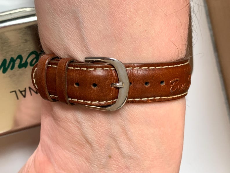 K&ouml;nnte beinhalten: Nahaufnahme eines braunen Leder-Uhrenarmbands mit wei&szlig;en N&auml;hten und einer silbernen Schnalle. Das Armband wird am Handgelenk getragen. Auf dem Uhrenarmband ist das Wort "Con" eingraviert.