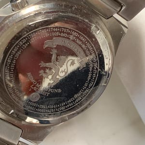 Puede incluir: Primer plano de la parte trasera de un reloj plateado. La esfera del reloj presenta texto grabado y un escudo con un le&oacute;n. El texto incluye las frases "IN DEFENS" y "NEMO ME IMPUNE LACESSIT".