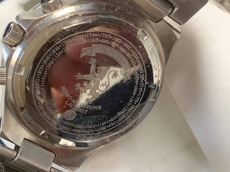 Puede incluir: Primer plano de la parte trasera de un reloj plateado. La esfera del reloj presenta texto grabado y un escudo con un le&oacute;n. El texto incluye las frases "IN DEFENS" y "NEMO ME IMPUNE LACESSIT".