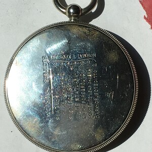 Op de afbeelding: Een zilverkleurig, cirkelvormig object met een ring en een kleine bal aan de bovenkant. Het object heeft ge&euml;tste tekst, waaronder "EXCEPTIONAL SUBJECTS" en "NORMAL EXPOSURE". De rand heeft een gedetailleerd, gestructureerd ontwerp.