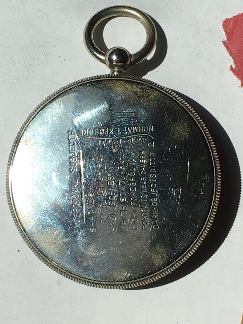 Op de afbeelding: Een zilverkleurig, cirkelvormig object met een ring en een kleine bal aan de bovenkant. Het object heeft ge&euml;tste tekst, waaronder "EXCEPTIONAL SUBJECTS" en "NORMAL EXPOSURE". De rand heeft een gedetailleerd, gestructureerd ontwerp.