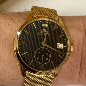 Peut inclure: Montre Astron dorée avec un cadran noir et des aiguilles dorées. La montre comporte un petit cadran de secondes, une fenêtre de date et un bracelet en maille. Le cadran a des rayures verticales et la marque "ASTRON" est affichée.
