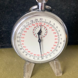 May include: A silver stopwatch with a white face and black hands. The stopwatch has a black ring around the edge with numbers from 1 to 60. The stopwatch is on a clear plastic stand.
