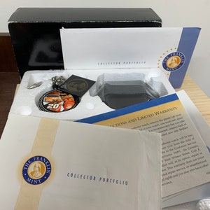 Op de afbeelding: Een zwarte doos met een wit interieur met daarin een zilveren zakhorloge met een zwarte wijzerplaat en een afbeelding van een raceautocoureur. De doos bevat ook een witte kaart met de tekst "COLLECTOR PORTFOLIO" en een klein kaartje met het logo "THE FRANKLIN MINT".