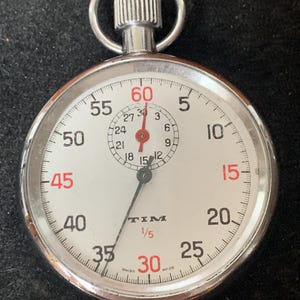 Peut inclure: Un chronomètre argenté avec un cadran blanc, des chiffres noirs et rouges. La montre a un petit cadran en haut et le mot "TIM" est visible. Le chronomètre est de conception classique, probablement utilisé pour chronométrer des événements.