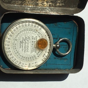 Op de afbeelding: Vintage Wynne's Infallible Belichtingsmeter in een metalen doos. De cirkelvormige meter heeft een glazen deksel en een kleine amberkleurige lens. De doos is open en onthult een blauwe kaart met tekst. Een fotografie-instrument.