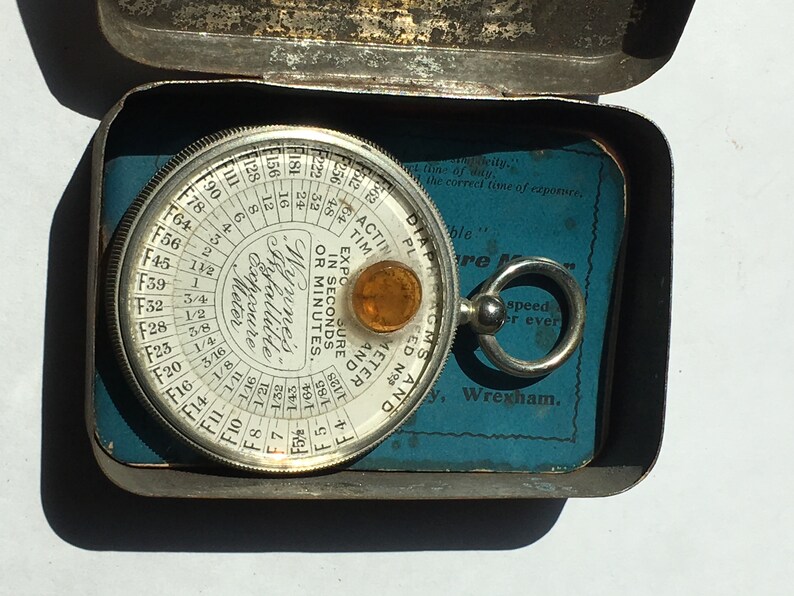 Op de afbeelding: Vintage Wynne's Infallible Belichtingsmeter in een metalen doos. De cirkelvormige meter heeft een glazen deksel en een kleine amberkleurige lens. De doos is open en onthult een blauwe kaart met tekst. Een fotografie-instrument.