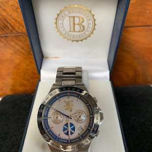 Puede incluir: Reloj de pulsera de acero inoxidable con esfera blanca, detalles azules y esfera detallada. El reloj est&aacute; en una caja azul con el logotipo "The Bradford Exchange". Cuenta con una pantalla de fecha y una correa de metal.