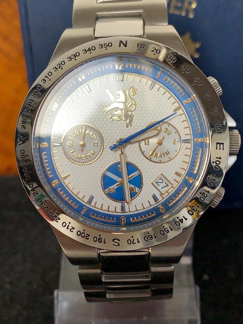 Puede incluir: Reloj de pulsera plateado con esfera blanca, detalles azules y detalles dorados. Cuenta con m&uacute;ltiples subesferas, una pantalla de fecha y el texto "SCOTLAND FOREVER" y "FRATH". La correa es de acero inoxidable.