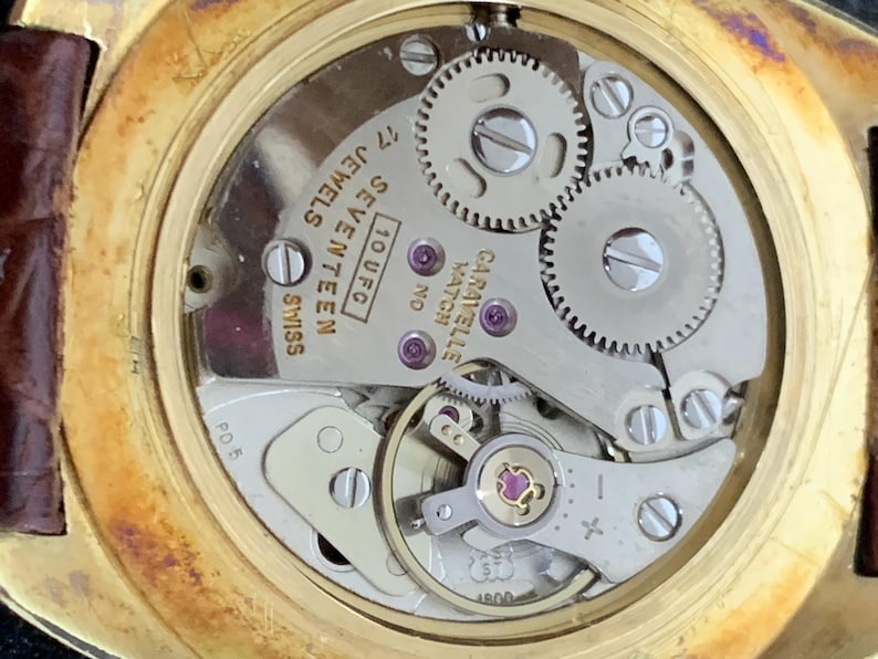 May include: Close-up of the back of a gold-tone wristwatch with a silver-tone face. The watch face has the text "10 UFC, SEVENTEEN JEWELS, SWISS" and "GARAVELLE WATCH NO."