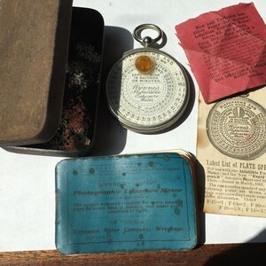 Op de afbeelding: Een verzameling vintage fotografische apparatuur, waaronder een ronde Wynne's Infallible Belichtingsmeter, een blauwe etui met tekst en een rood papier met tekst. De meter heeft een zilveren behuizing en een gele kern. De items zijn gerangschikt op een witte ondergrond.