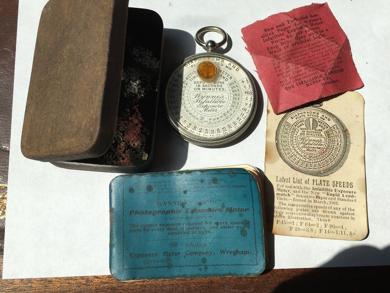 Op de afbeelding: Een verzameling vintage fotografische apparatuur, waaronder een ronde Wynne's Infallible Belichtingsmeter, een blauwe etui met tekst en een rood papier met tekst. De meter heeft een zilveren behuizing en een gele kern. De items zijn gerangschikt op een witte ondergrond.