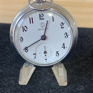 Peut inclure: Une montre de poche argentée avec un cadran blanc et des aiguilles noires. La montre porte une inscription noire qui dit "SERVICES" et est posée sur un support en plastique transparent.