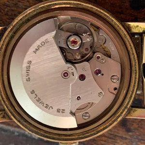 May include: Close-up of a vintage watch movement. The watch mechanism is visible, showcasing intricate gears, jewels, and the words "Swiss Made" and "25 Jewels". The watch has a gold-toned case and a "Fixo-Flex" band.