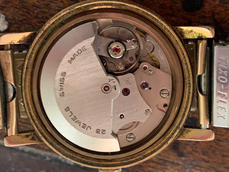 May include: Close-up of a vintage watch movement. The watch mechanism is visible, showcasing intricate gears, jewels, and the words "Swiss Made" and "25 Jewels". The watch has a gold-toned case and a "Fixo-Flex" band.