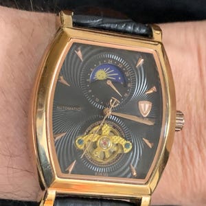 Pode incluir: Um relógio dourado com mostrador preto e pulseira de couro preta. O relógio possui uma complicação de fase lunar e um design de coração aberto. O mostrador do relógio também tem o texto "AUTOMATIC" e "V-TIME".