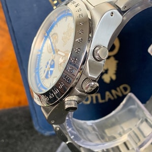 Puede incluir: Reloj de pulsera plateado con esfera azul y plateada. El reloj tiene una correa de acero inoxidable y una esfera detallada con m&uacute;ltiples subesferas y marcas. El reloj se muestra sobre un soporte transparente, con una caja azul en el fondo.