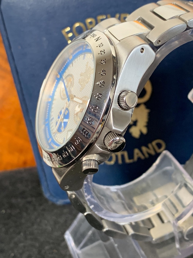 Puede incluir: Reloj de pulsera plateado con esfera azul y plateada. El reloj tiene una correa de acero inoxidable y una esfera detallada con m&uacute;ltiples subesferas y marcas. El reloj se muestra sobre un soporte transparente, con una caja azul en el fondo.