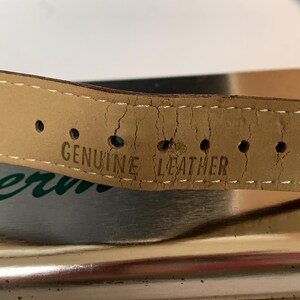 K&ouml;nnte beinhalten: Nahaufnahme eines hellbraunen Lederarmbands mit dem Stempel "GENUINE LEATHER". Das Armband weist Gebrauchsspuren auf, mit Rissen und L&ouml;chern f&uuml;r die Schnalle. Wei&szlig;e N&auml;hte sind an den R&auml;ndern sichtbar.