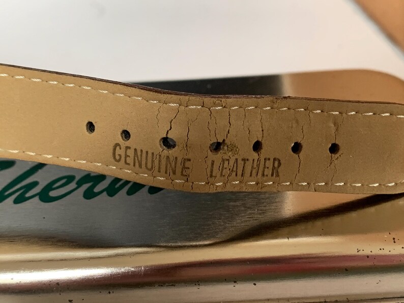 K&ouml;nnte beinhalten: Nahaufnahme eines hellbraunen Lederarmbands mit dem Stempel "GENUINE LEATHER". Das Armband weist Gebrauchsspuren auf, mit Rissen und L&ouml;chern f&uuml;r die Schnalle. Wei&szlig;e N&auml;hte sind an den R&auml;ndern sichtbar.