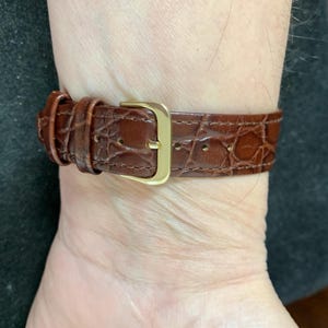 May include: A brown leather watch strap with a gold buckle. The strap has a crocodile pattern.