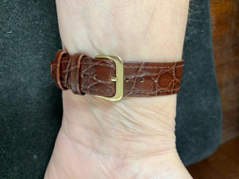 May include: A brown leather watch strap with a gold buckle. The strap has a crocodile pattern.