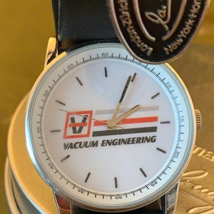 May include: A silver wristwatch with a white face displaying the text "VACUUM ENGINEERING" and a logo. The watch has a black leather strap and gold hands. The watch face has a minute track around the edge.