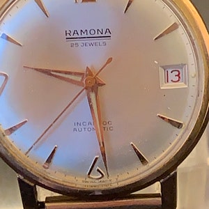 May include: Close-up of a vintage Ramona wristwatch. The watch features a silver face with gold-tone hands and markers. The text "RAMONA" and "25 JEWELS" are visible. The date is displayed as "13". The watch has a gold-tone case and a brown metal band.