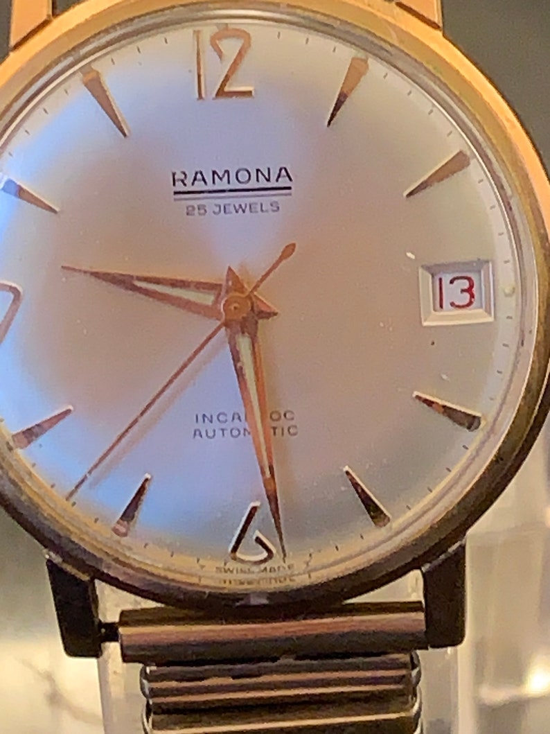 May include: Close-up of a vintage Ramona wristwatch. The watch features a silver face with gold-tone hands and markers. The text "RAMONA" and "25 JEWELS" are visible. The date is displayed as "13". The watch has a gold-tone case and a brown metal band.