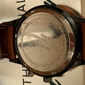 K&ouml;nnte beinhalten: Nahaufnahme einer Ben Sherman-Uhr mit braunem Lederarmband. Das Zifferblatt der Uhr ist schwarz mit einem silberfarbenen R&uuml;cken. Auf der R&uuml;ckseite der Uhr steht "LICENCE FROM SHERMAN C.I.N.L. THE ORIGINAL WATER RESISTANT 50M".