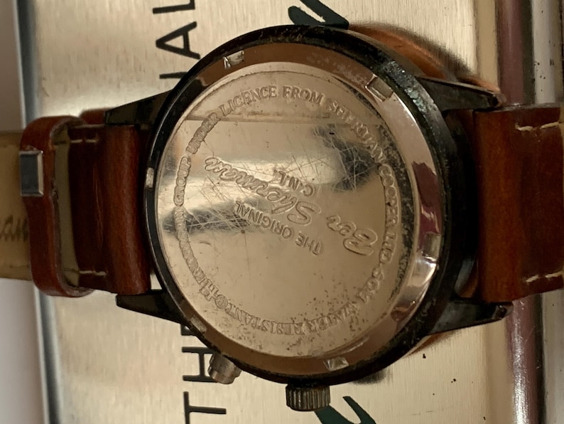 K&ouml;nnte beinhalten: Nahaufnahme einer Ben Sherman-Uhr mit braunem Lederarmband. Das Zifferblatt der Uhr ist schwarz mit einem silberfarbenen R&uuml;cken. Auf der R&uuml;ckseite der Uhr steht "LICENCE FROM SHERMAN C.I.N.L. THE ORIGINAL WATER RESISTANT 50M".